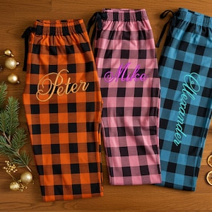 May include: Three pairs of plaid pajama pants in orange, pink, and blue. Each pair has a name embroidered on the front: Peter, Mike, and Alexander. The pants are arranged on a wooden surface with holiday decorations.