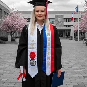 May include: A graduate in a black cap and gown, wearing a white, red, and blue nursing sash. The sash reads "NURSING" and "BSN RN". She holds a diploma with a red ribbon and a blue folder. The background features a building and pink flowering trees.