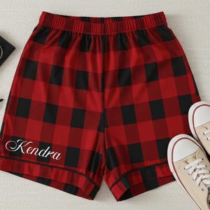 May include: Red and black plaid pajama shorts with the name "Kendra" in white script. The shorts are paired with beige canvas sneakers and sunglasses.
