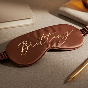 May include: A brown satin sleep mask with the name "Brittany" in gold script. The mask has an elastic band and is lying on a textured surface next to a gold pen and a closed book.