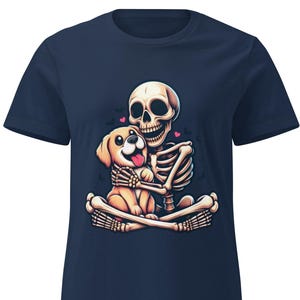 Könnte beinhalten: Marineblaues T-Shirt mit einer Cartoon-Illustration eines Skeletts, das einen Golden Retriever-Welpen umarmt. Das Design umfasst rosa Herzen und Schmetterlinge. Das Bild ist mittig auf der Vorderseite des T-Shirts platziert.