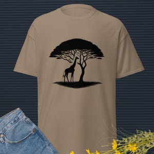 Könnte beinhalten: Ein beiges T-Shirt mit einer schwarzen Silhouette einer Giraffe unter einem Baum. Der Baum hat ein breites, abgerundetes Blätterdach. Das Shirt wird mit blauen Jeans und gelben Blumen gezeigt.