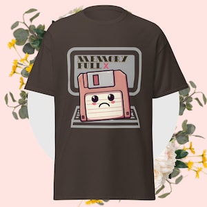 Könnte beinhalten: Braunes T-Shirt mit einem Cartoon-Design einer Diskette mit einem traurigen Gesicht auf einem Computerbildschirm. Der Text "MEMORY FULL X" befindet sich über der Diskette. Das Design verwendet Rosa-, Grau- und Brauntöne.