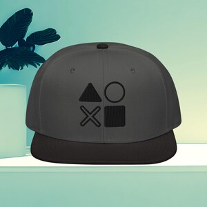 Könnte beinhalten: Grau-schwarze Baseballkappe mit einem schwarzen, gestickten Design aus Dreieck, Kreis, X und Quadrat. Die Kappe hat einen flachen Schirm und eine strukturierte Krone, geeignet für Freizeitkleidung.
