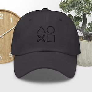 Può includere: Un cappellino da baseball nero con i simboli dei pulsanti del controller PlayStation: triangolo, cerchio, X e quadrato. Il cappello è realizzato in un materiale morbido e ha una visiera curva. Il cappello è di colore nero.