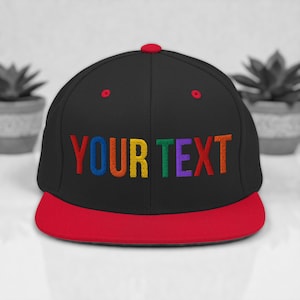May include: Black baseball cap with a red brim and button. The cap features the embroidered text "YOUR TEXT" in rainbow colors. The cap is a flat-brim style.