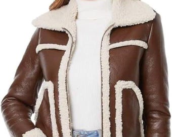 May include: A brown faux leather jacket with a cream-colored shearling lining and trim. The jacket features a zip-up front, a wide collar, and pockets. The model is wearing blue jeans and a white turtleneck. The jacket is a stylish outerwear option.