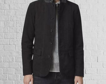 Puede incluir: Un blazer negro con cuello de cuero, cierre de botones y dos bolsillos delanteros. La chaqueta se lleva sobre un suéter gris y vaqueros oscuros. El fondo es una pared de ladrillo blanco.