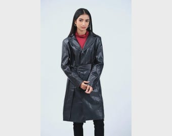Pode incluir: Um trench coat longo de couro preto com cinto na cintura e gola de lapela clássica. O casaco é usado sobre uma gola alta vermelha. A modelo está usando calças pretas. O casaco parece ser na altura do joelho.