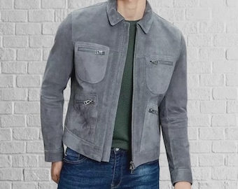 Può includere: Giacca in camoscio grigio con collo e multiple tasche con cerniera. La giacca ha una chiusura frontale con cerniera e viene abbinata a jeans blu. Il modello indossa una camicia verde sotto.