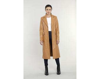 Pode incluir: Um trench coat comprido, de cor bege, com gola de lapela clássica e corte sob medida. O casaco tem fecho de botões e bolsos. A modelo está vestida com o casaco com um top branco, jeans azul escuro e botas pretas.
