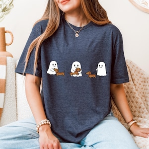 May include: Dark blue t-shirt featuring three white ghost figures with dachshund dogs. One ghost holds a dog, another walks a dog on a leash, and the third ghost is petting a dog. The shirt is a casual style.