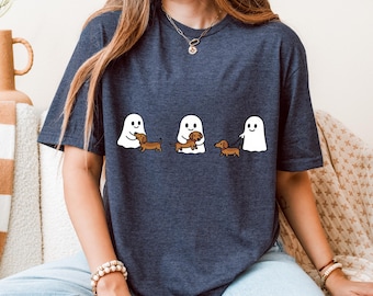 May include: Dark blue t-shirt featuring three white ghost figures with dachshund dogs. One ghost holds a dog, another walks a dog on a leash, and the third ghost is petting a dog. The shirt is a casual style.