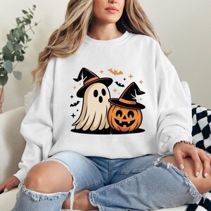 May include: White sweatshirt with a Halloween graphic. The design features a cartoon ghost and a jack-o'-lantern, both wearing witch hats. Bats and stars are included, creating a festive Halloween theme.