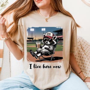 Può includere: Una t-shirt beige a maniche corte con la grafica di un procione che indossa occhiali da sole e un berretto con la scritta RAISING BALLERS, seduto su una sedia pieghevole su un campo da baseball con una bevanda. Testo I live here now sotto.