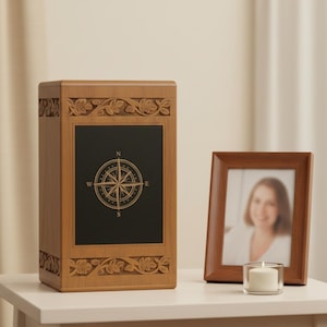 May include: A wooden urn with a compass rose design on a black panel. The urn is light brown with carved floral details. A framed photo and a lit candle sit nearby on a white table.