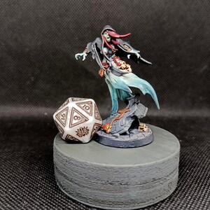 Op de afbeelding: Een miniatuur fantasy figuur van een spectrale tovenares met rood haar en een vloeiende blauwe jurk, staand op een rotsachtige basis. Daarnaast een grote, witte, veelzijdige dobbelsteen met zwarte markeringen. De figuur en de dobbelsteen staan op een grijs, cilindrisch platform.