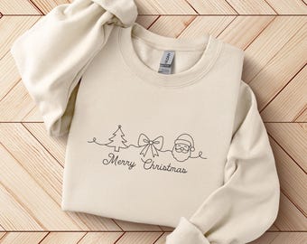 May include: Cream-colored sweatshirt with rolled-up sleeves. The front features a black line drawing of a Christmas tree, bow, Santa Claus, and the words "Merry Christmas". The sweatshirt is laid on a light brown wooden surface.