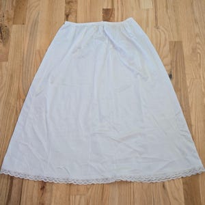 May include: A white slip skirt with a lace trim at the hem. The skirt is made of a lightweight fabric and is laid out on a wooden surface. The skirt is a solid white color.