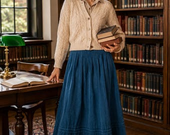 Könnte beinhalten: Eine beige Strickjacke mit Zopfmuster und Knöpfen über einem gestreiften Hemd mit Kragen, kombiniert mit einem langen blauen Faltenrock und dunkelbraunen Schnürstiefeln. Das Outfit wird durch eine runde Brille und drei gehaltene Bücher ergänzt.