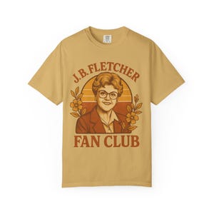 Puede incluir: Camiseta de color amarillo mostaza con un diseño retro que presenta un retrato de una mujer con gafas y una blazer marrón. El texto dice "J.B. FLETCHER FAN CLUB" encima y debajo del retrato, con elementos florales.