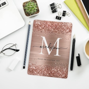 May include: A rose gold notebook with the name "Madison" and the letter "M" in white. The notebook has a glittery border and the text "Name or Custom Text" at the bottom. Other items on the white surface include a pencil, pen, and a small potted cactus.
