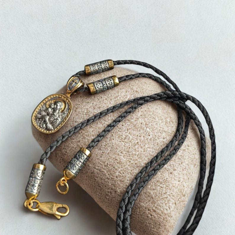 May include: A silver and gold pendant necklace featuring an oval pendant with a religious figure. The pendant hangs from a black braided cord with silver and gold accents. The necklace is displayed on a beige surface.