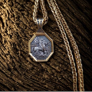 May include: A gold-toned pendant necklace with an octagonal pendant featuring a detailed relief of a knight on horseback slaying a dragon. The pendant is attached to a gold chain, and the background is a textured wooden surface.