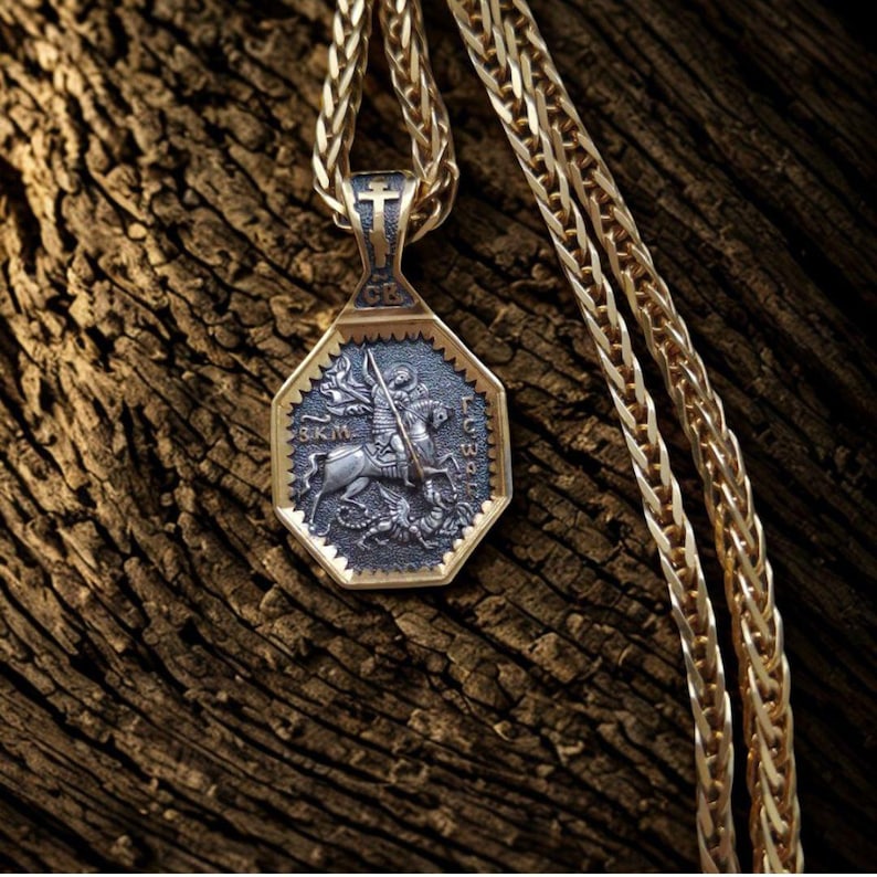 May include: A gold-toned pendant necklace with an octagonal pendant featuring a detailed relief of a knight on horseback slaying a dragon. The pendant is attached to a gold chain, and the background is a textured wooden surface.