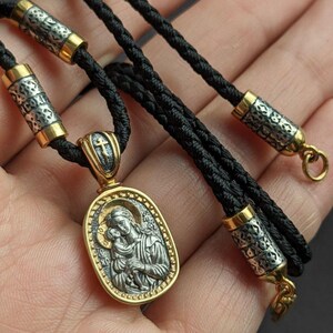 May include: A gold and silver pendant necklace featuring an oval pendant with a religious icon of a woman and child. The pendant hangs on a black cord with silver and gold accents. The necklace is approximately 50 cm long.