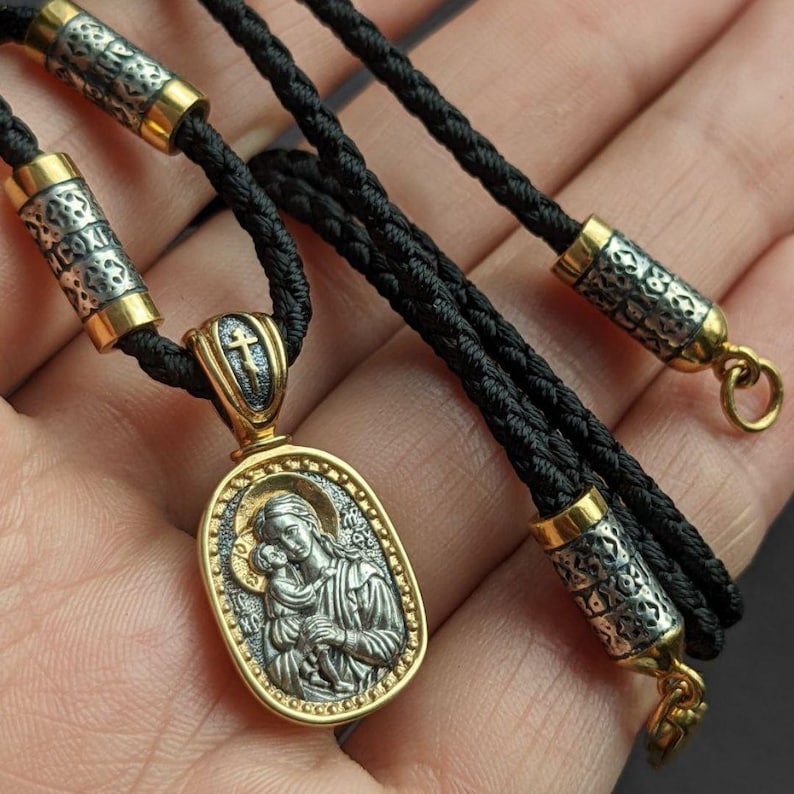 May include: A gold and silver pendant necklace featuring an oval pendant with a religious icon of a woman and child. The pendant hangs on a black cord with silver and gold accents. The necklace is approximately 50 cm long.