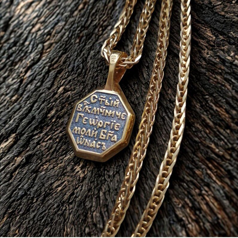 May include: A gold-colored pendant necklace with an octagonal charm. The charm has blue text and a gold border. The chain is a wheat chain style. The necklace is displayed on a dark wood surface.