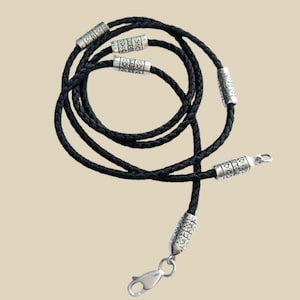 May include: Black braided leather necklace with silver-colored decorative beads and a clasp. The necklace is coiled and lies against a neutral background. The beads have a detailed pattern.