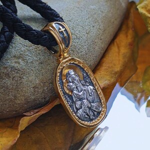 May include: An oval gold-coloured pendant with a religious icon, depicting a figure holding a child. The pendant has a cross detail and is attached to a black cord. The design features silver and gold accents. The pendant is likely made of metal.