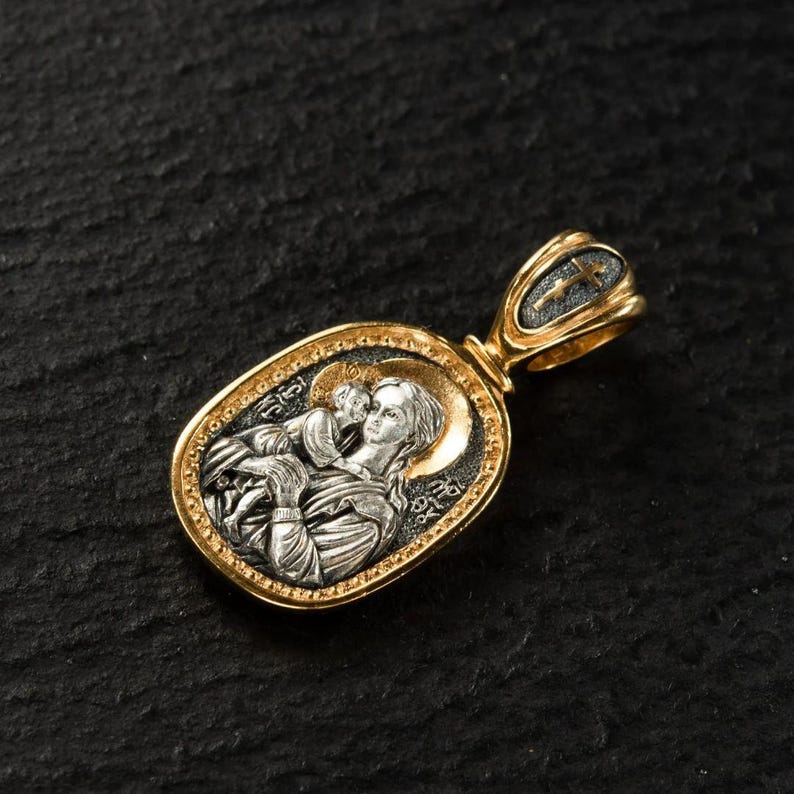 May include: An oval pendant with a religious icon design. The pendant has a gold-coloured bail with a cross, and a silver-coloured oval frame with a detailed image of a woman and child. The pendant is set against a dark background.