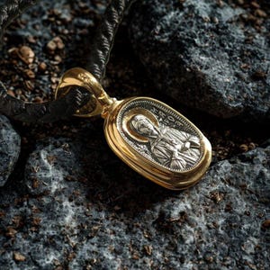 May include: Gold pendant featuring an oval, silver religious icon, suspended from a black, braided cord. The pendant is set against a backdrop of dark grey rocks. The contrast highlights the shiny metal against the rough stone.