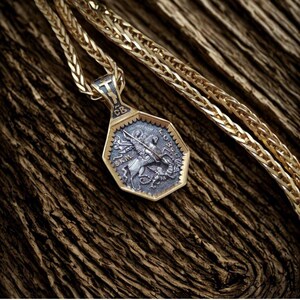 May include: Gold-toned pendant necklace with a detailed, octagonal pendant. The pendant features a silver-colored relief of a knight on horseback slaying a dragon. The chain is a wheat style, and the background is a textured, brown surface.