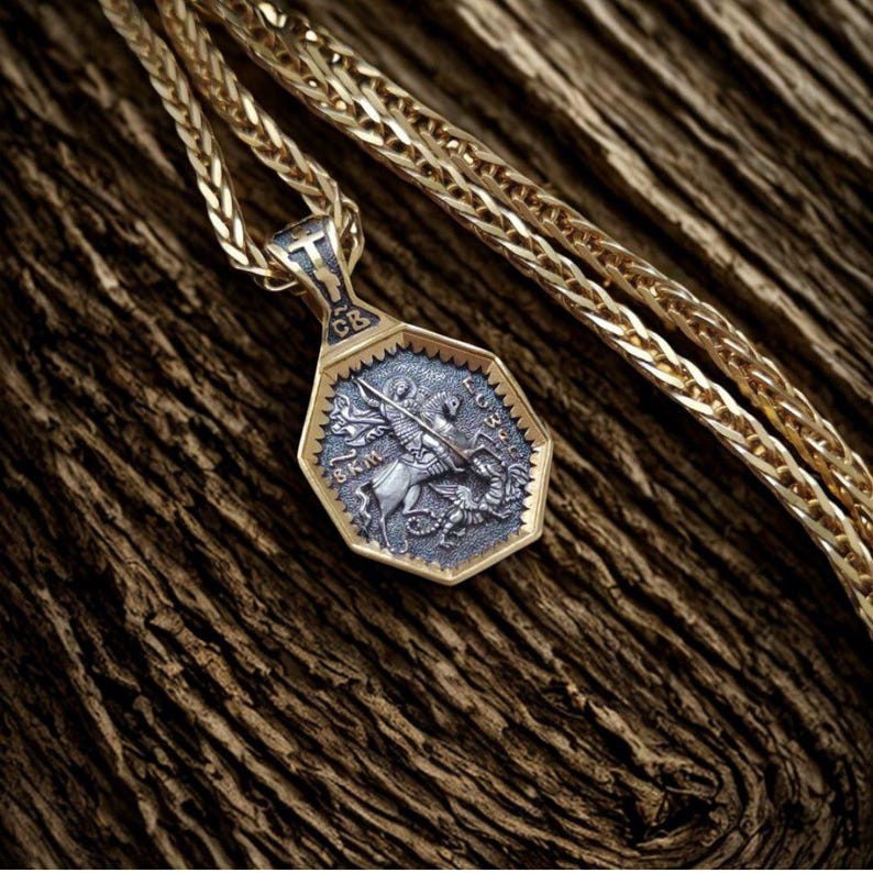 May include: Gold-toned pendant necklace with a detailed, octagonal pendant. The pendant features a silver-colored relief of a knight on horseback slaying a dragon. The chain is a wheat style, and the background is a textured, brown surface.