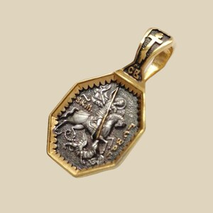 May include: A gold-toned pendant with a detailed silver-colored central design. The pendant features a depiction of a figure on horseback spearing a creature, with text and a cross. The bail has a cross design.