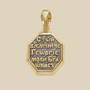 May include: Gold-toned pendant with an octagonal face and a loop for a chain. The pendant features Cyrillic text in a contrasting dark color, framed by a gold border. The pendant is likely made of gold.