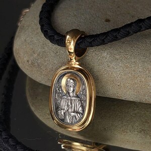 May include: A gold pendant with an oval silver religious icon, depicting a haloed figure. The pendant is suspended from a black woven cord. The icon is detailed with religious imagery. The pendant is set against a neutral stone background.