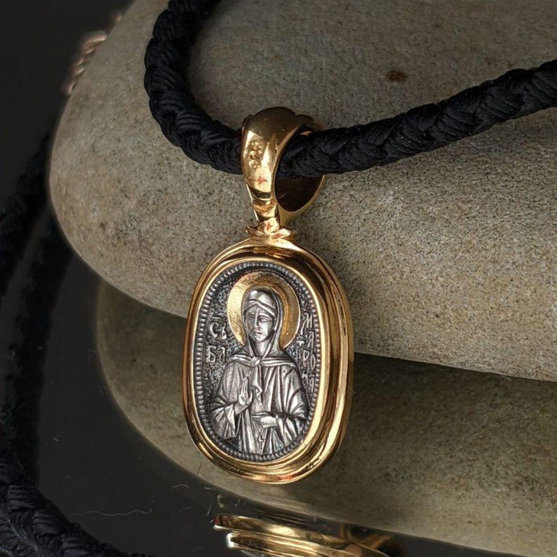 May include: A gold pendant with an oval silver religious icon, depicting a haloed figure. The pendant is suspended from a black woven cord. The icon is detailed with religious imagery. The pendant is set against a neutral stone background.