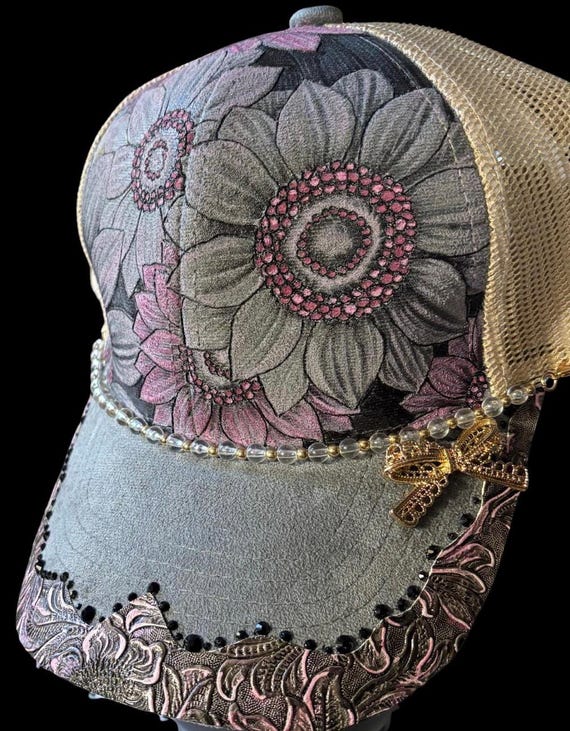 Pink sunflowers hand-burned using a hand held wood burner. Pink accents and charms. Faux grey suede with tan mesh ponytail back.