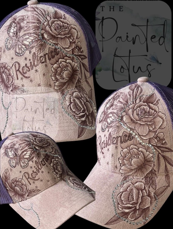 Lavender faux suede ponytail cap, hand-burned, with a wood burner, flowers and the wording “Resilience”. Trailing rhinestones.