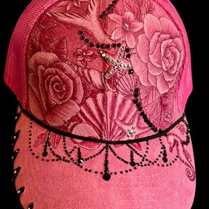 May include: Pink trucker hat with a floral design. The hat features a mesh back and a curved brim. The front panel is adorned with embossed roses, seashells, and a starfish, embellished with black beads and rhinestones.