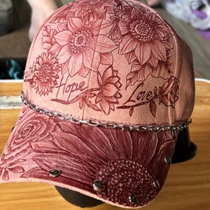 May include: A peach-colored baseball cap featuring an embossed floral pattern of sunflowers and roses. The words "Hope" and "Love" are scripted across the front panels. A silver-toned metal chain rests above the brim, which is accented with small silver studs.
