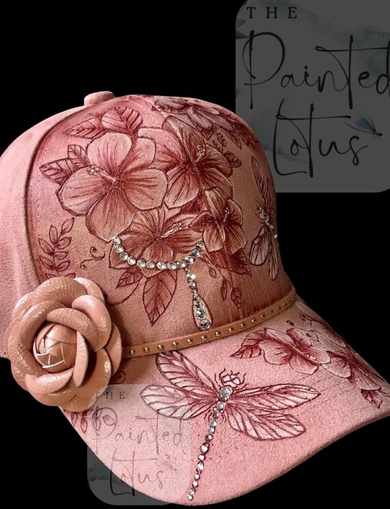 Pink faux suede ball cap, hand-burned, with a wood burner, flowers and dragonflies.Rhinestones, trailing rhinestones, and studded lacing.