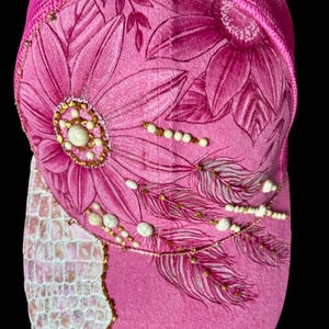 May include: A bright pink trucker hat featuring a mesh back and a front panel decorated with hand-painted floral designs in darker pink tones. The cap is embellished with white beads, gold-toned accents, and a textured white faux-reptile skin patch.