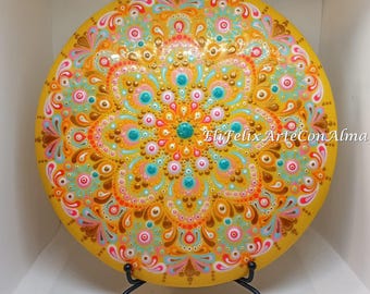 Puede incluir: Un plato decorativo redondo amarillo con un diseño de mandala detallado. El mandala presenta un patrón simétrico con acentos turquesa, rosa y dorado. El plato está sostenido por un soporte de metal negro. El texto en el plato dice "EliFelix ArteConAlma".