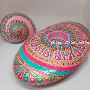 May include: Two hand-painted decorative stones. One is a spiral shape, the other is oval with a circular indentation. Both are painted in pink, teal, and gold with intricate dot patterns. The oval stone has a teal and pink stripe.